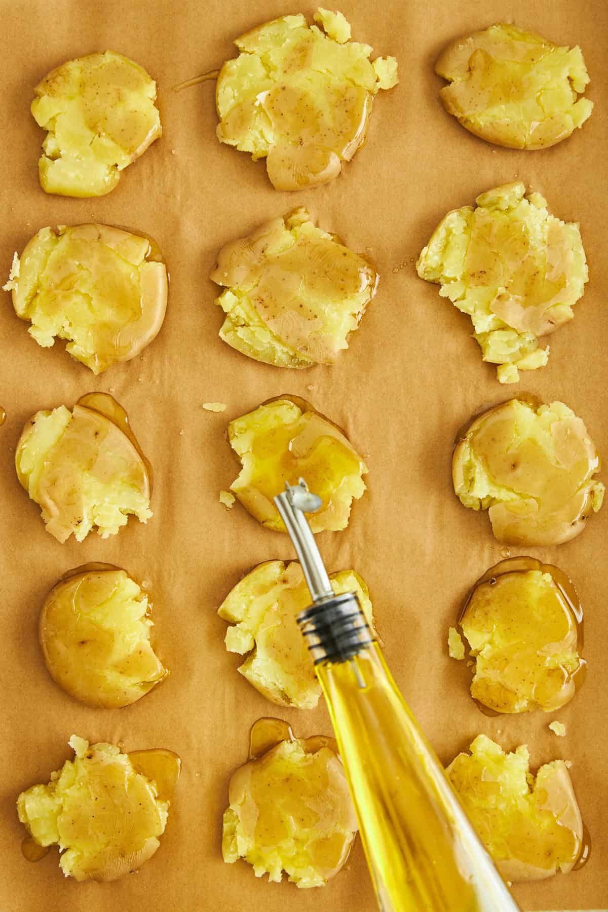 Oil being drizzled over uncooked smashed potatoes on a parchment lined baking sheet.