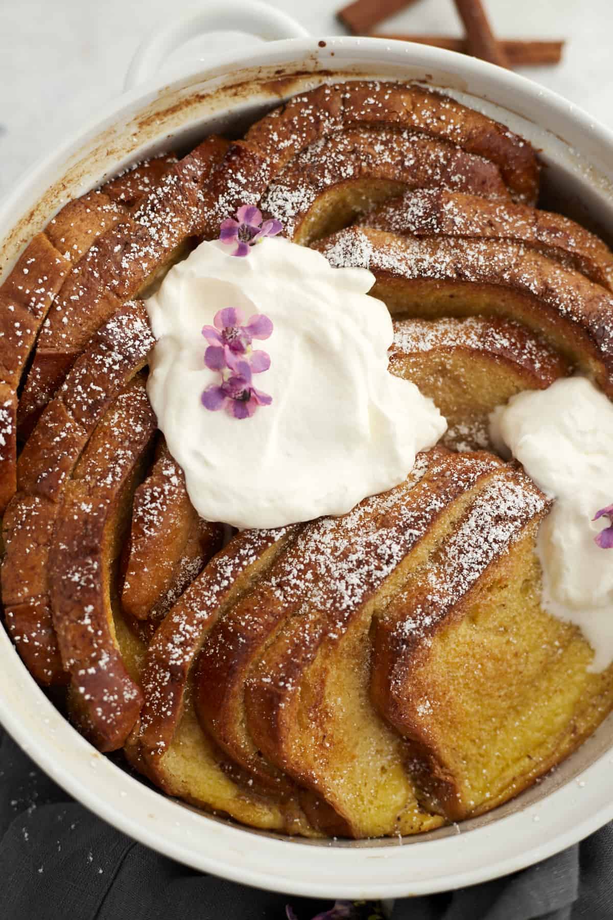 closeup of a white baking dish with baked cinnamon french toast topped with powdered sugar and marscapone cheese