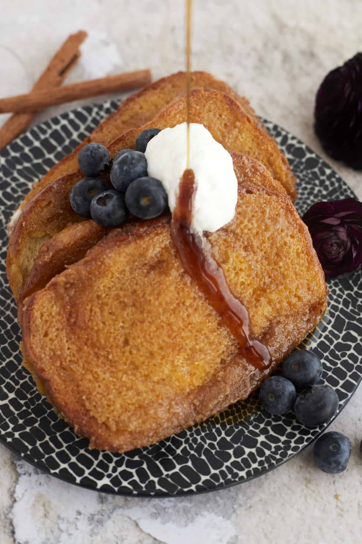 a plate of easy french toast with caramel sauce, whipped cream, and blueberries with syrup being poured on top 
