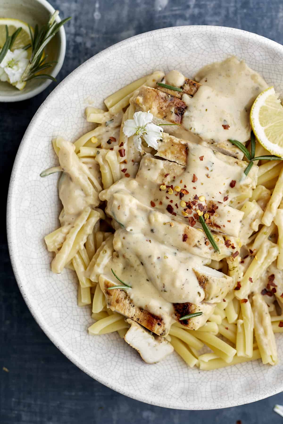 overhead image of creamy herb chicken and pasta noodles on a white plate topped with red pepper flakes, lemon wedges, and fresh herbs