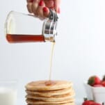 a stack of cottage cheese protein pancakes recipe on a white plate with blueberries on the side, strawberries in the background, and a woman's hand pouring syrup on top