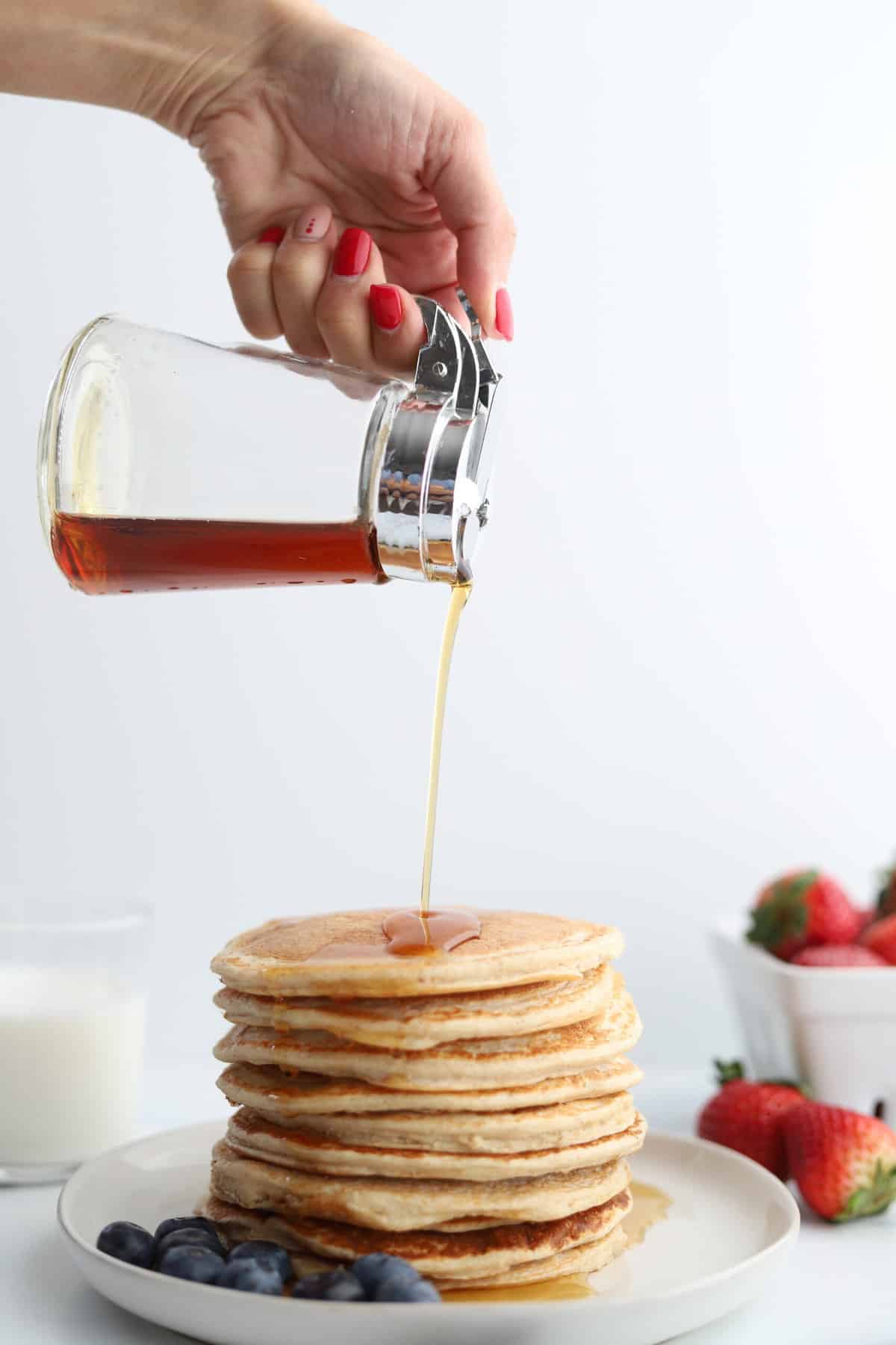 A stack of cottage cheese protein pancakes recipe on a white plate with blueberries on the side, strawberries in the background, and a woman's hand pouring syrup on top.
