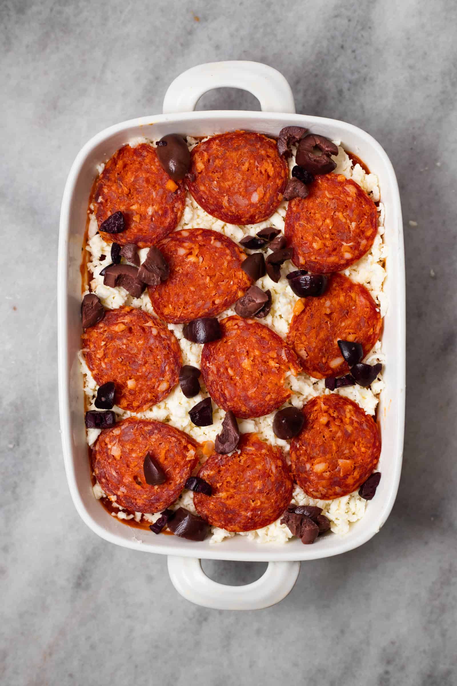 pepperoni and olives spread over mozzarella cheese, marinara sauce, and cheese spread in a baking dish