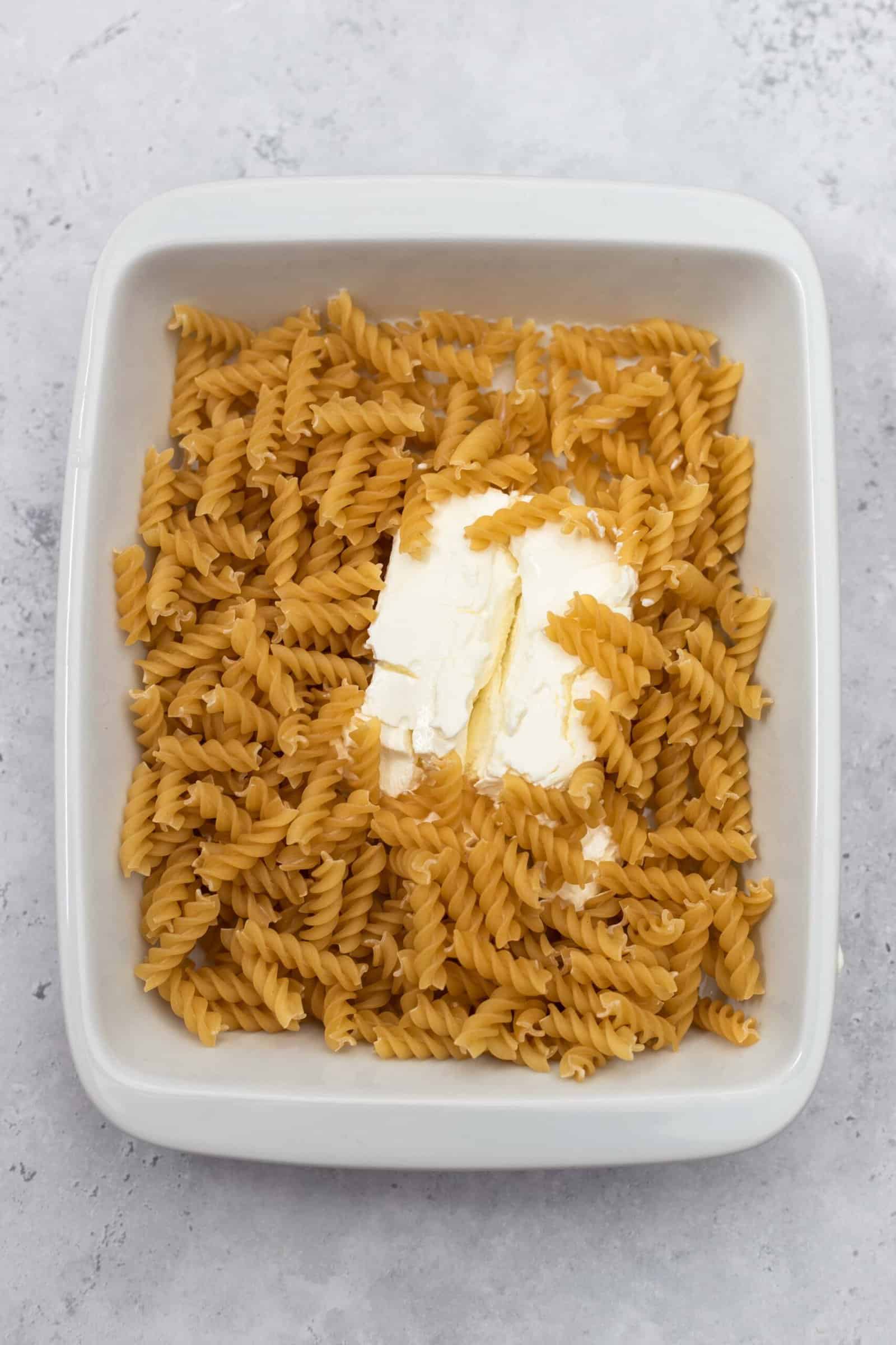 a block of cream cheese in the center of a baking dish surrounded by raw corkscrew pasta