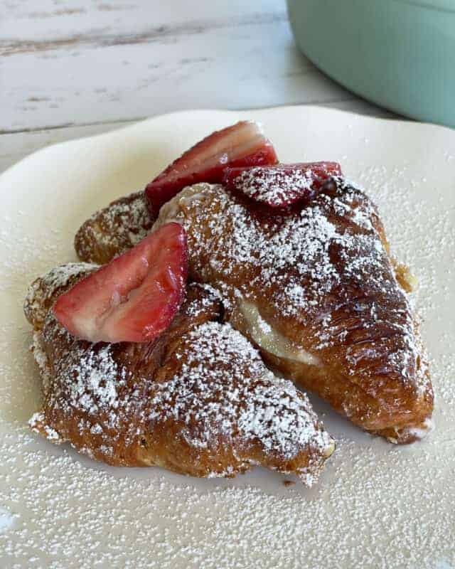 two strawberry cheesecake french toast croissants on a plate topped with powdered sugar and sliced strwaberries