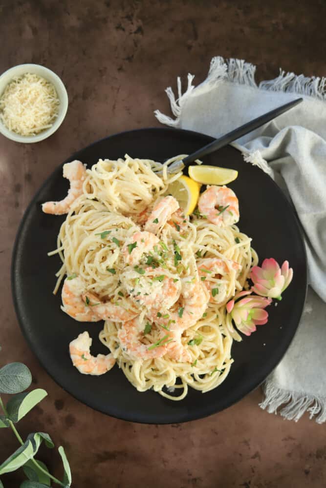 overhead image of a black place with baked garlic shrimp scampi topped with fresh parsley and lemon wedges and two flowers for garnish on the side of the plate and a fork twisted around the noodles and a small white bowl with Parmesan and a gray towel off to the side