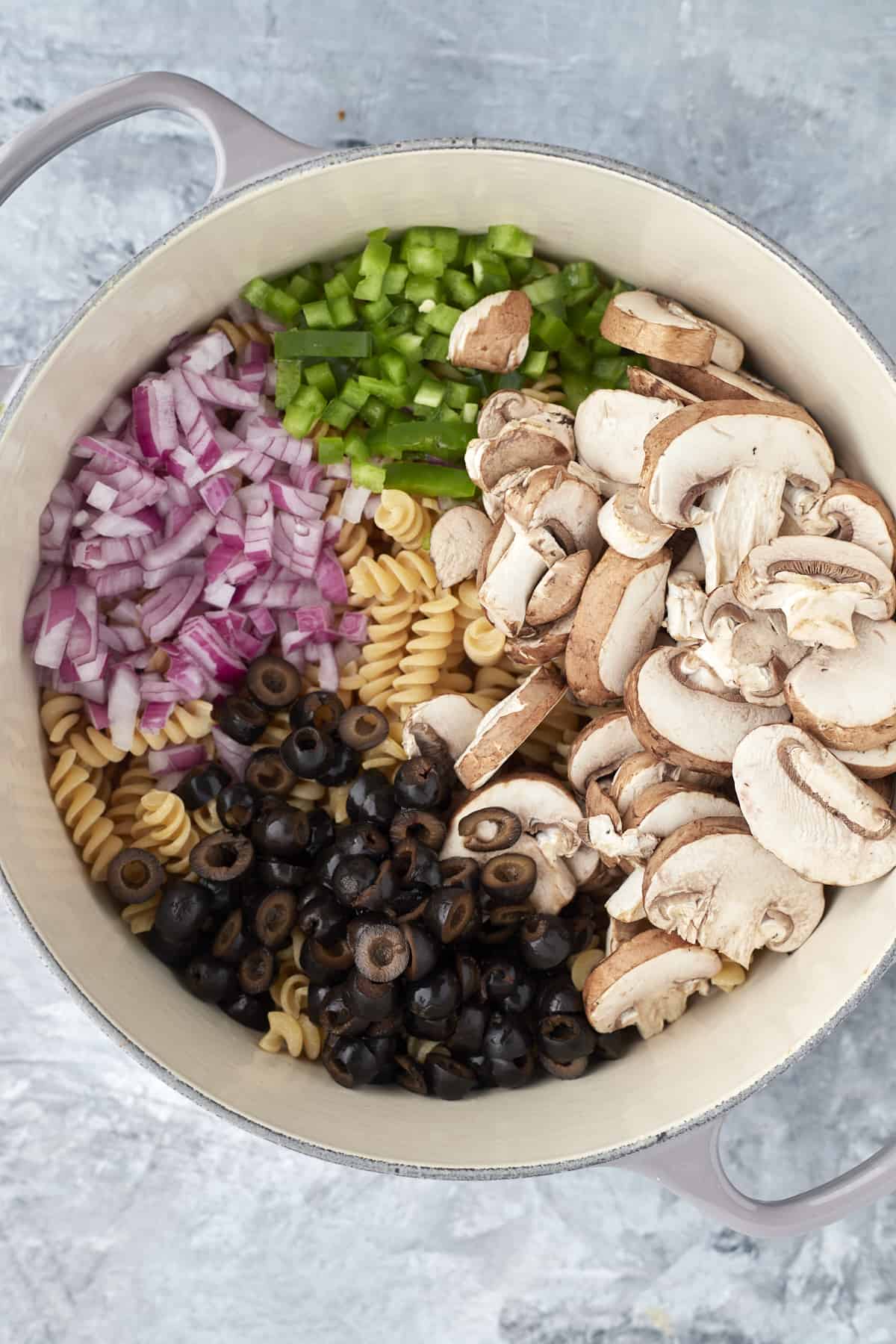 overhead image of a pot full of uncooked pasta, red onions, green peppers, mushrooms, and black olives