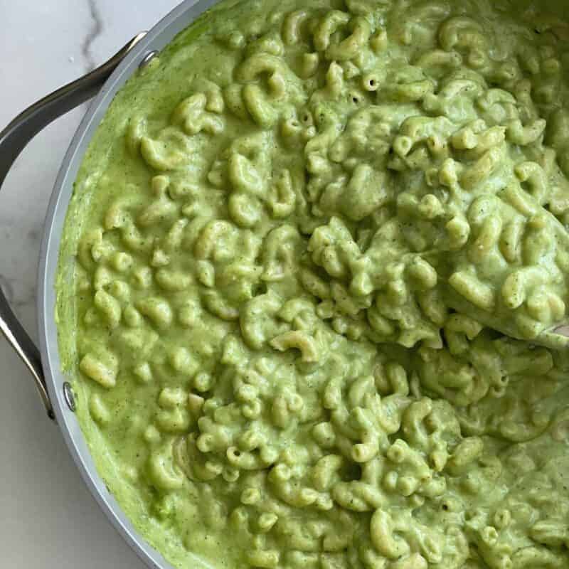 A pot of green mac and cheese being stirred with a wooden spoon.