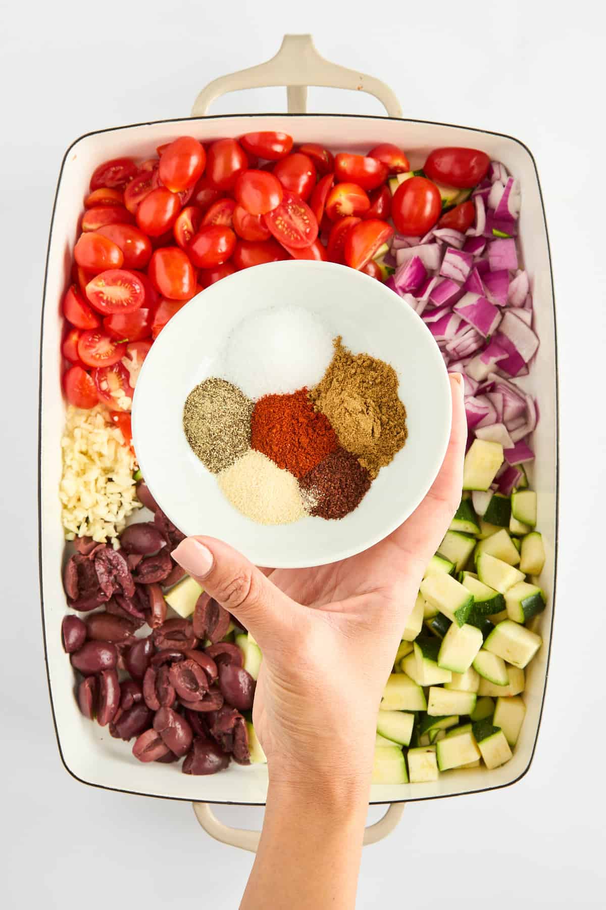 Spices being sprinkled over tomatoes, red onions, zucchini, olives, and garlic in a baking dish. 