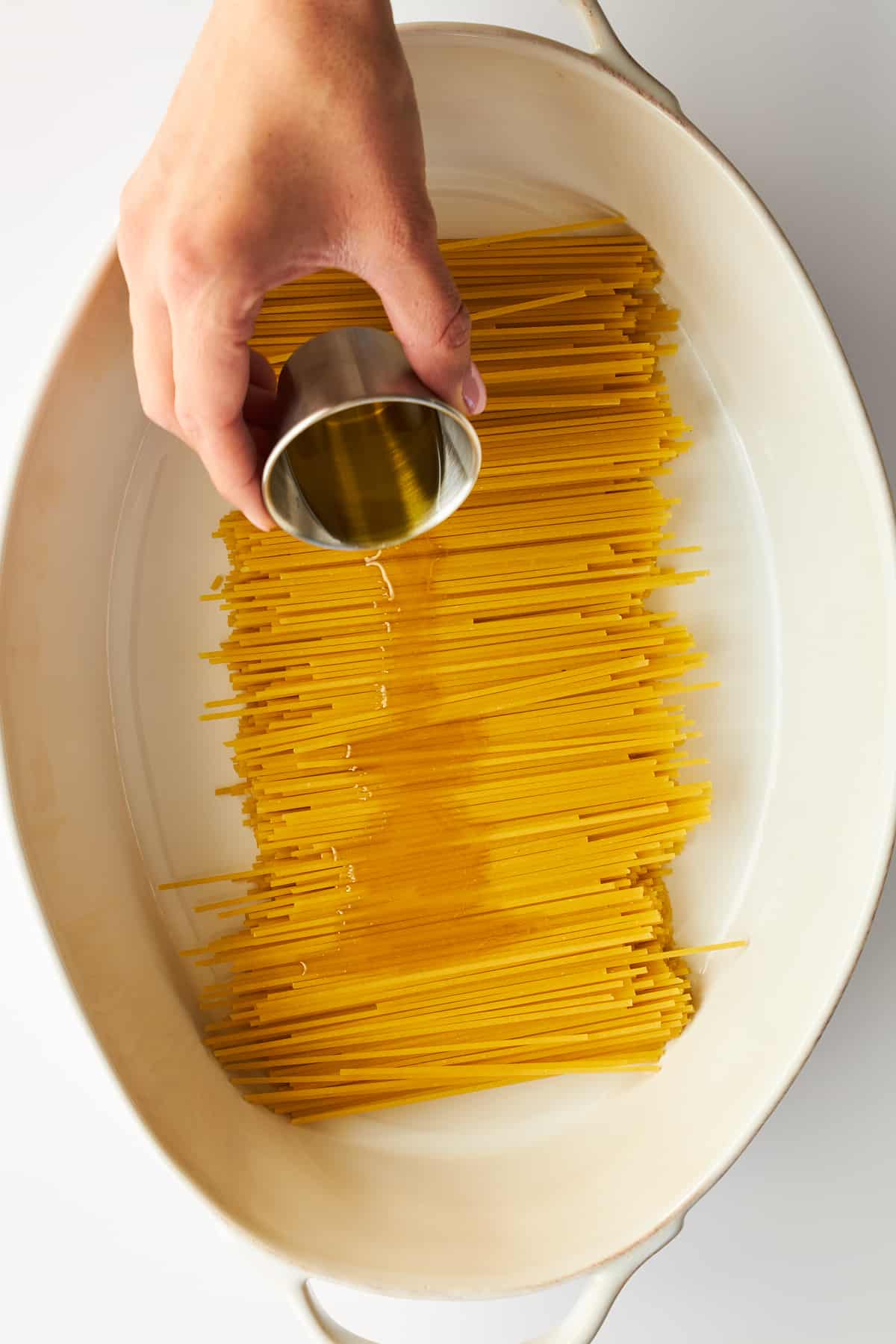 Olive oil being poured over spaghetti noodles in an oval baking dish.