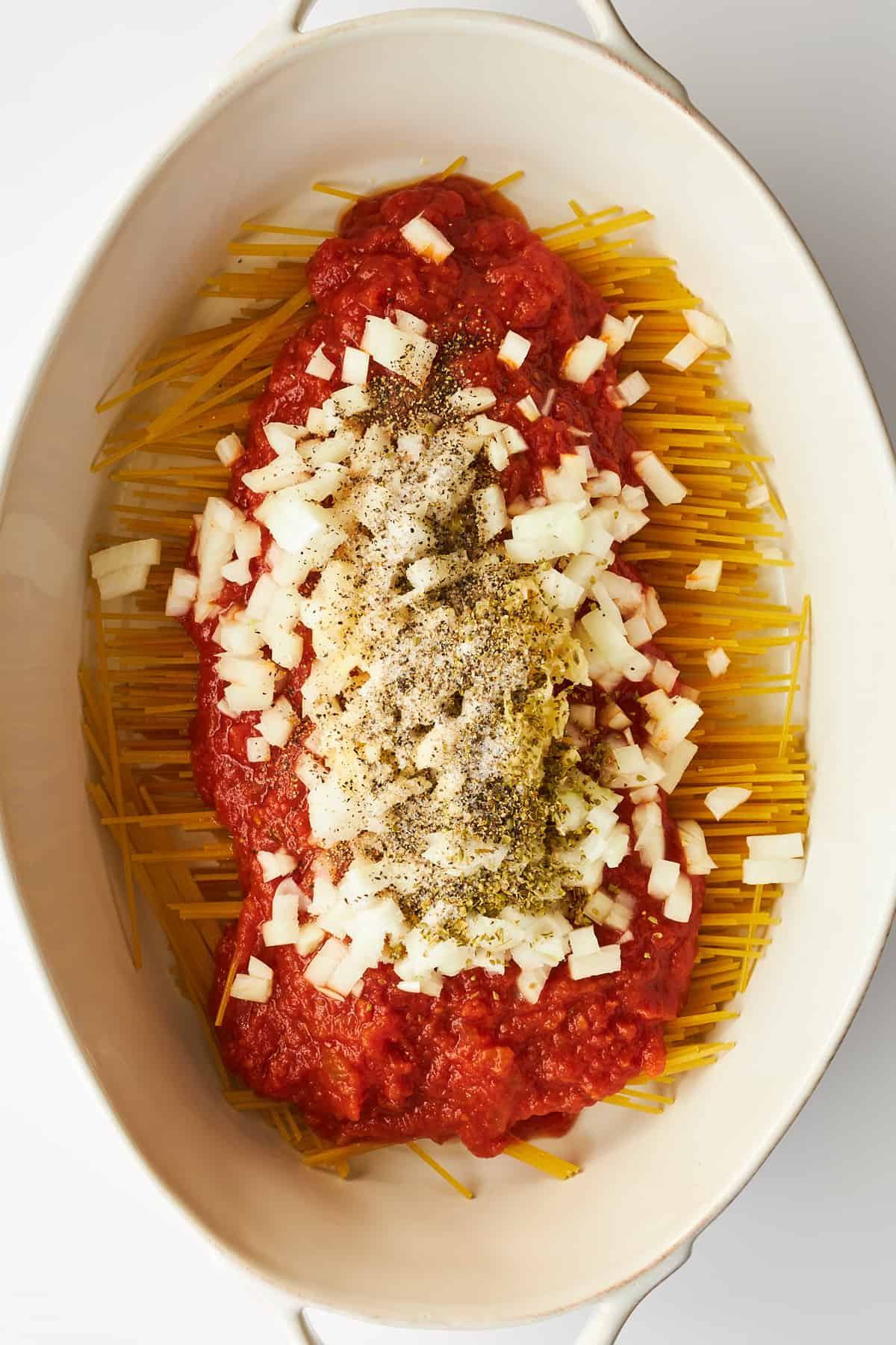 Onions and seasonings on top of marinara sauce covering spaghetti noodles in an oval baking dish.