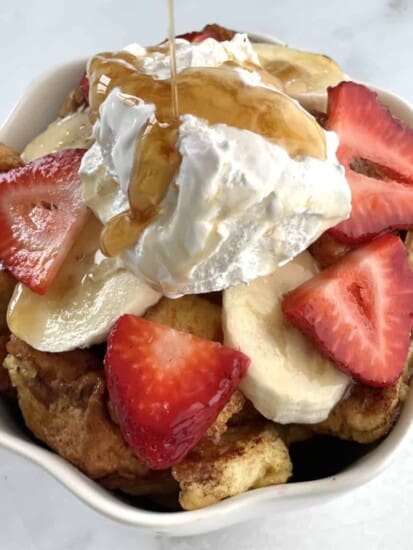 French toast cereal with banana coins, strawberry slices, whipped cream, and maple syrup on top.