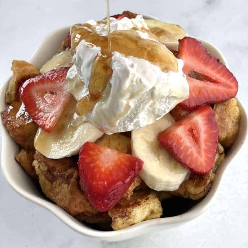 French toast cereal with banana coins, strawberry slices, whipped cream, and maple syrup on top.