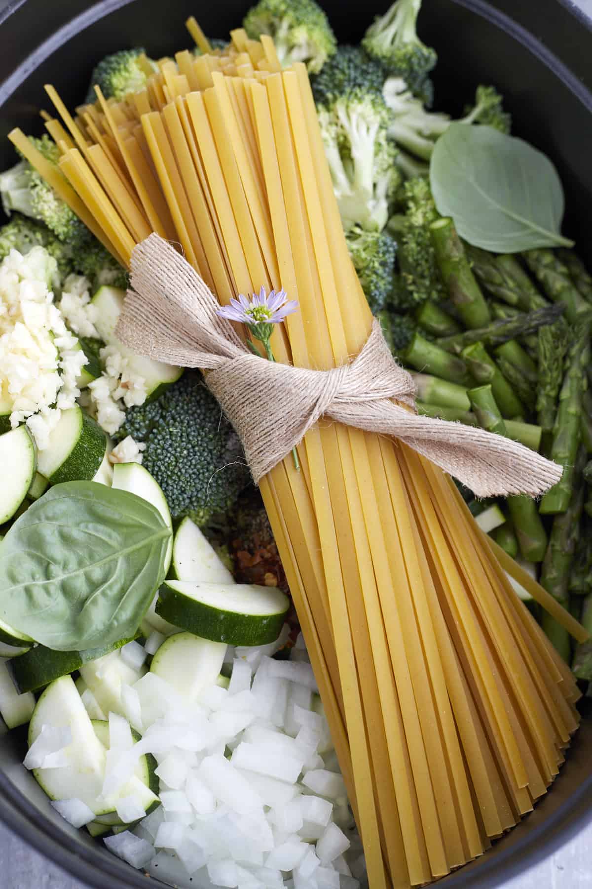 overhead image of a bundle of pasta noodles over onions, zucchini, garlic, herbs, broccoli, and asparagus