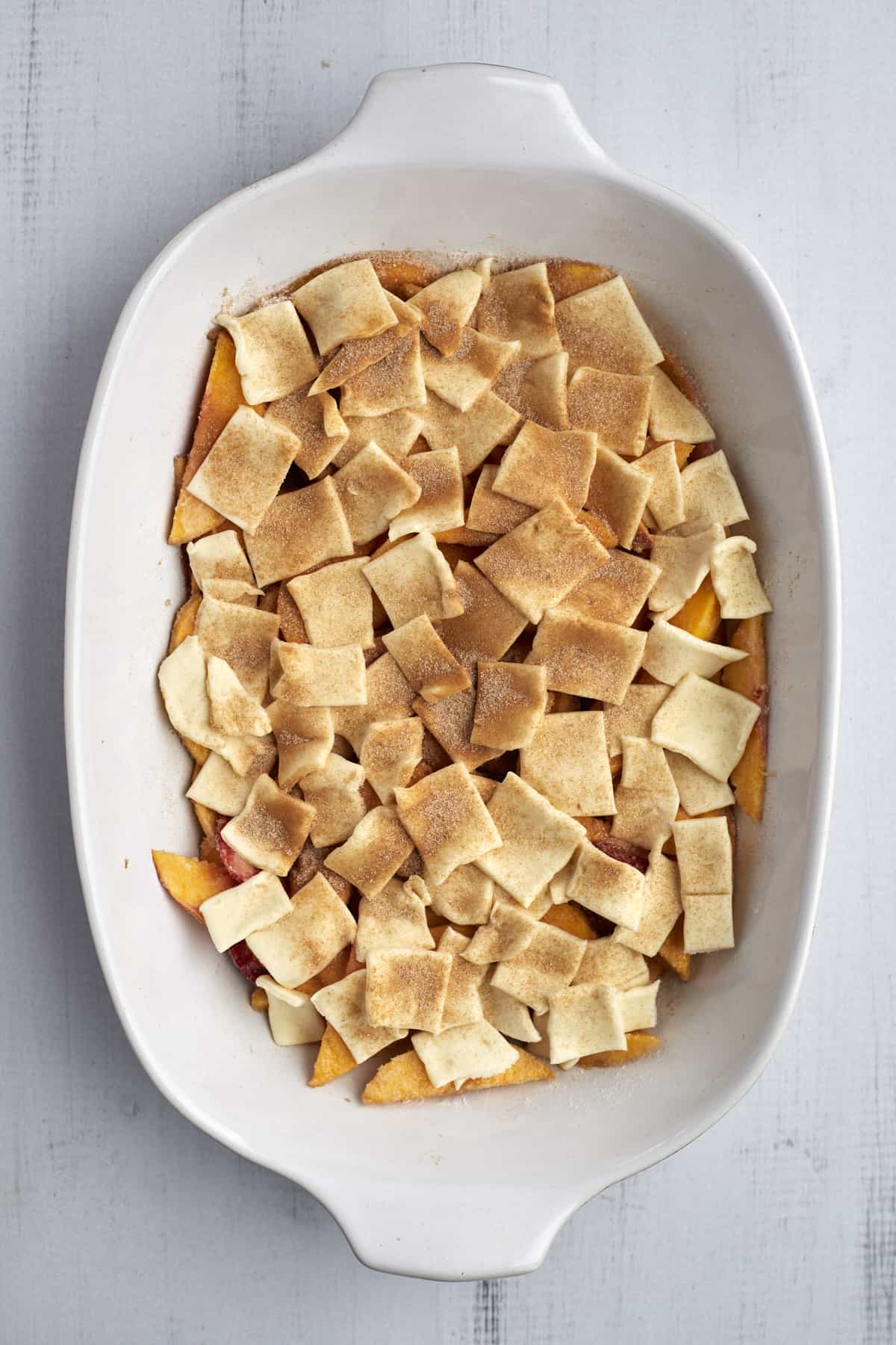 peaches and strawberries in a baking dish topped with raw dough pieces, sugar, and cinnamon