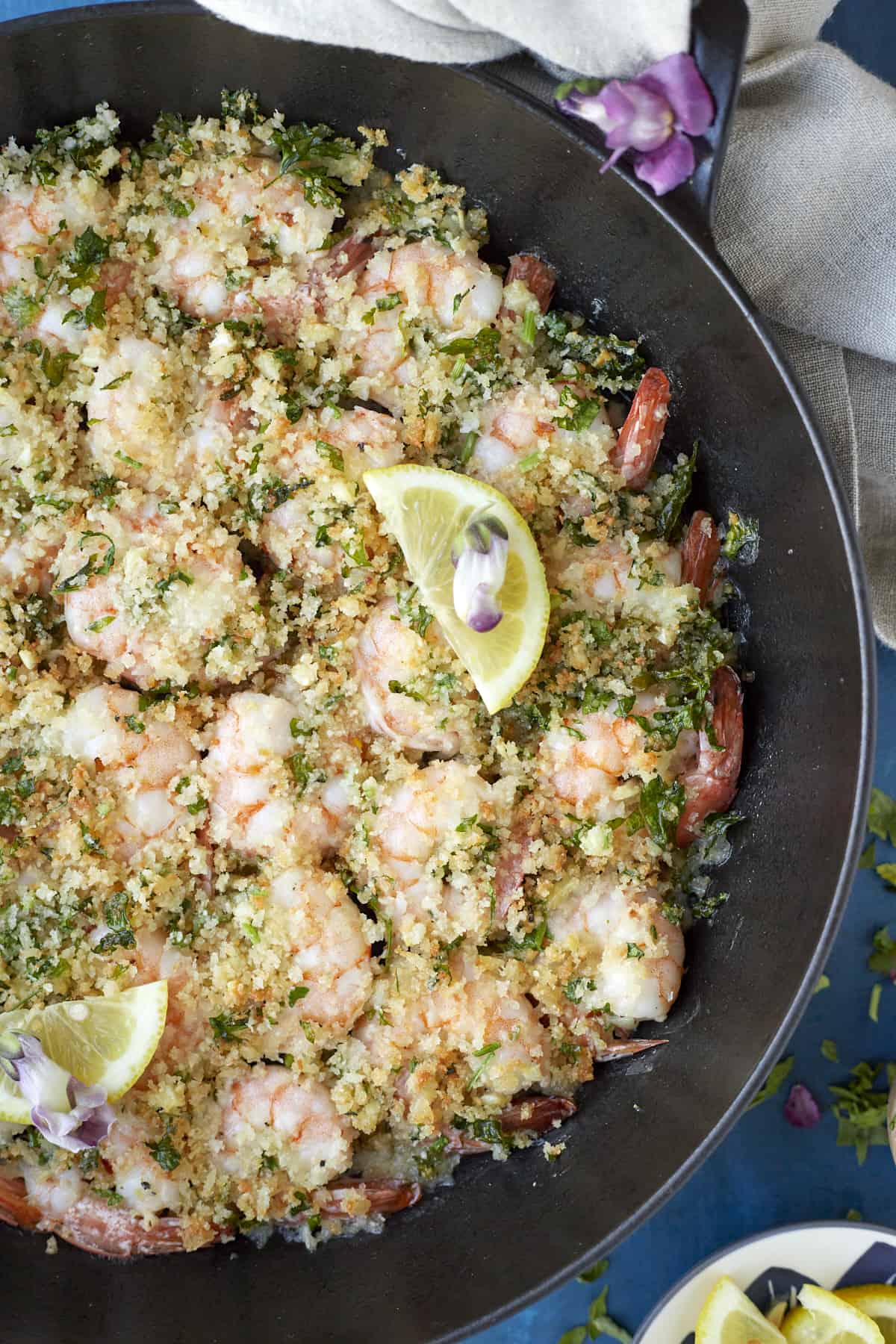 overhead image of baked shrimp scampi in a skillet topped with fresh herbs and a lemon wedge