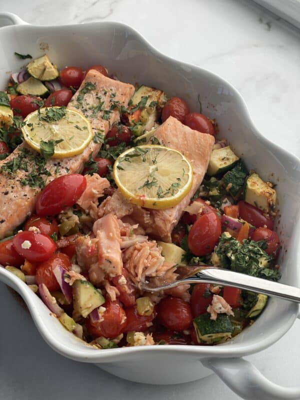 baked feta salmon with veggies in a baking dish topped with lemon slices being cut with a fork