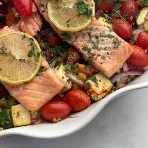close up image of baked feta salmon with veggies in a white baking dish topped with lemon slices and fresh herbs