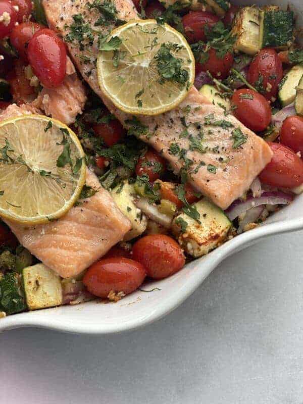 close up image of baked feta salmon with veggies in a white baking dish topped with lemon slices and fresh herbs