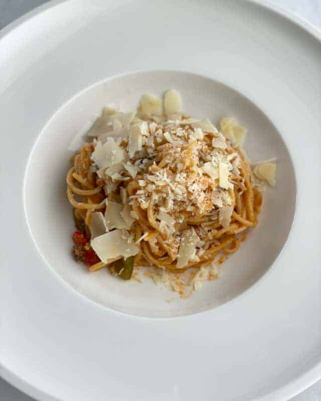 A plate with a serving of baked spaghetti bolognese recipe topped with shaved parmesan.