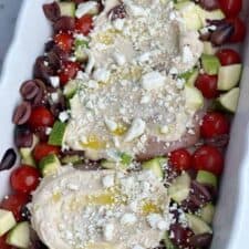 a baking dish with three raw chicken breasts topped with hummus, cheese, and oil surrounded by vegetables