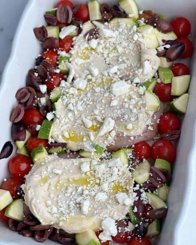 a baking dish with three raw chicken breasts topped with hummus, cheese, and oil surrounded by vegetables