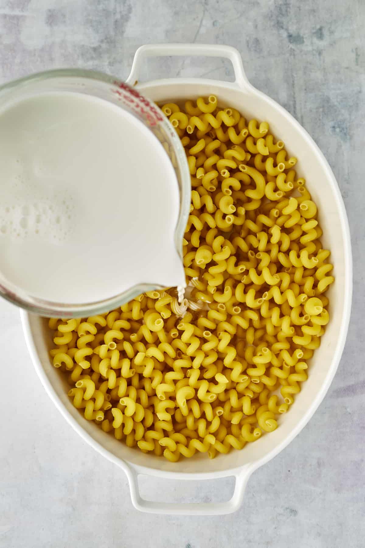 milk being poured over cavatappi noodles in an oval baking dish.