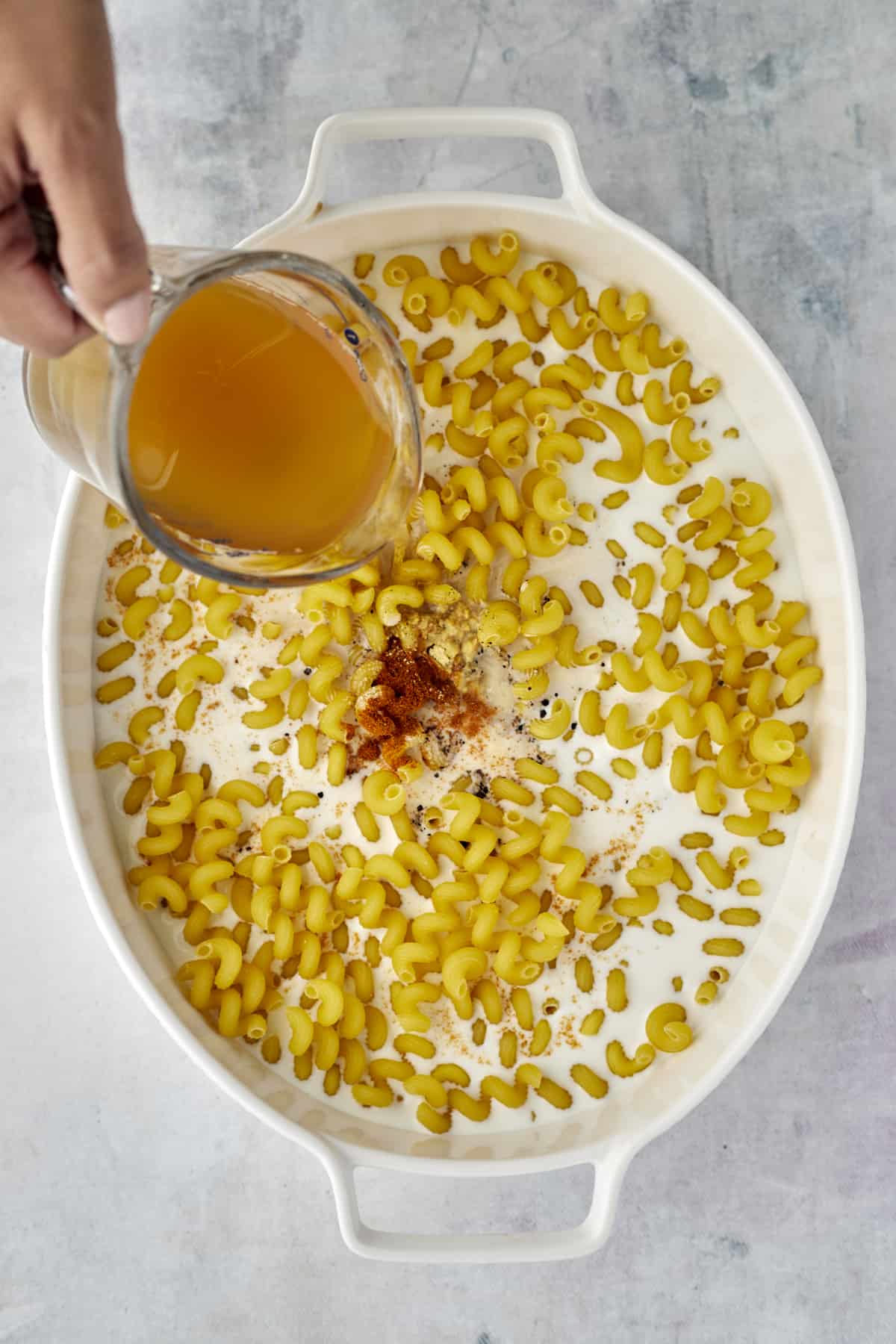 broth and spices being poured over milk and cavatappi noodles in an oval baking dish 