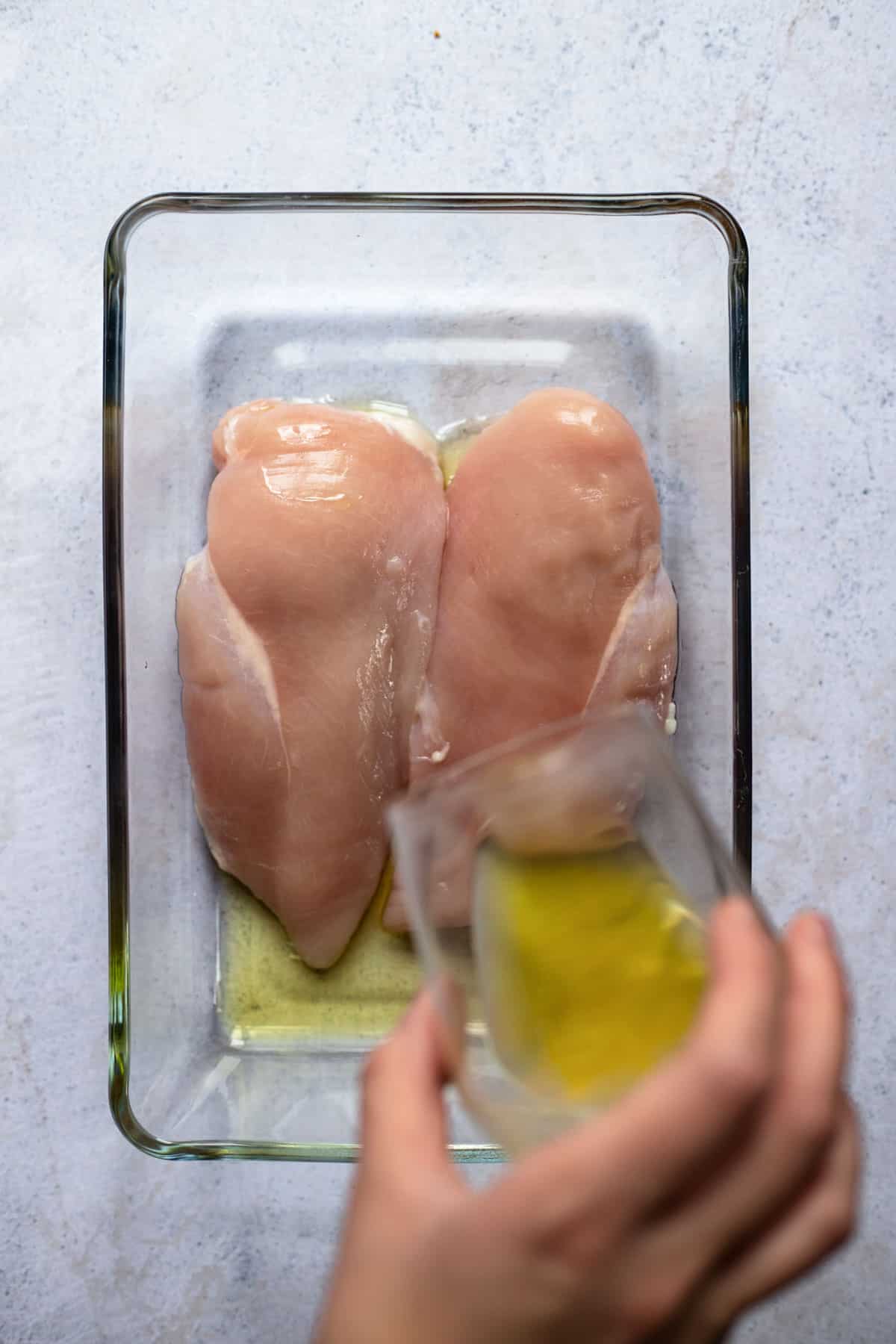 two raw chicken breasts in a baking dish with oil being poured over them 