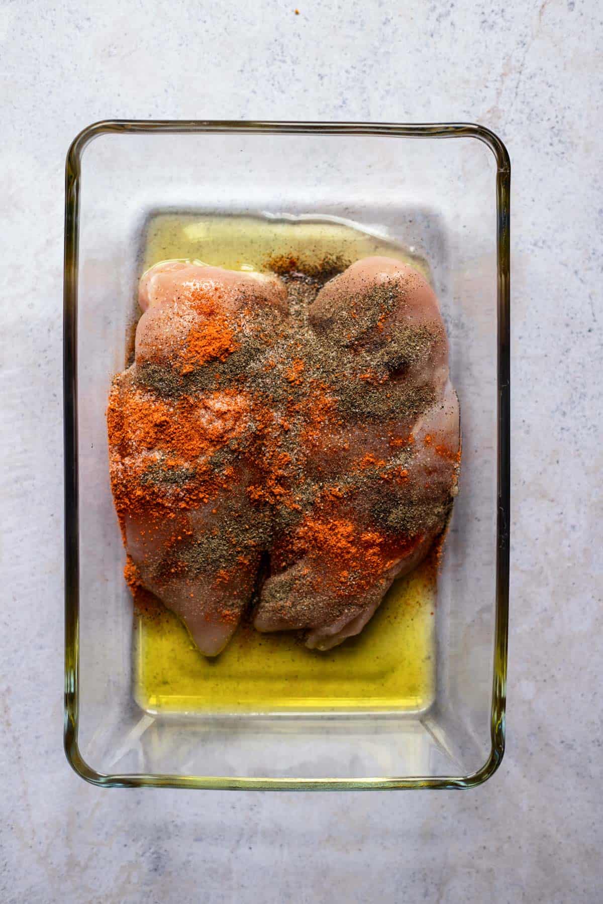 two raw chicken breasts in a baking dish topped with oil and spices
