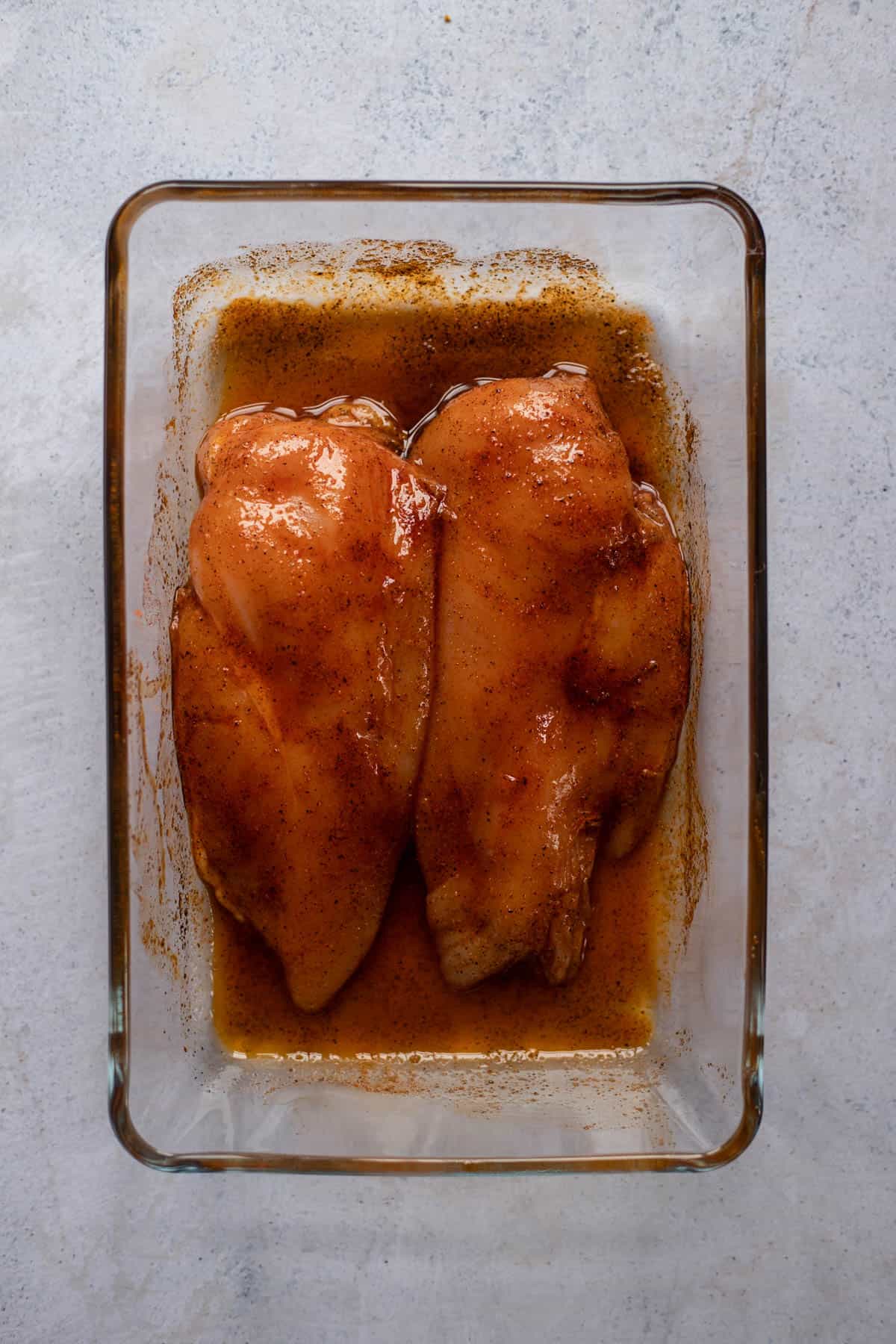 two raw chicken breasts in a baking dish rubbed with seasonings