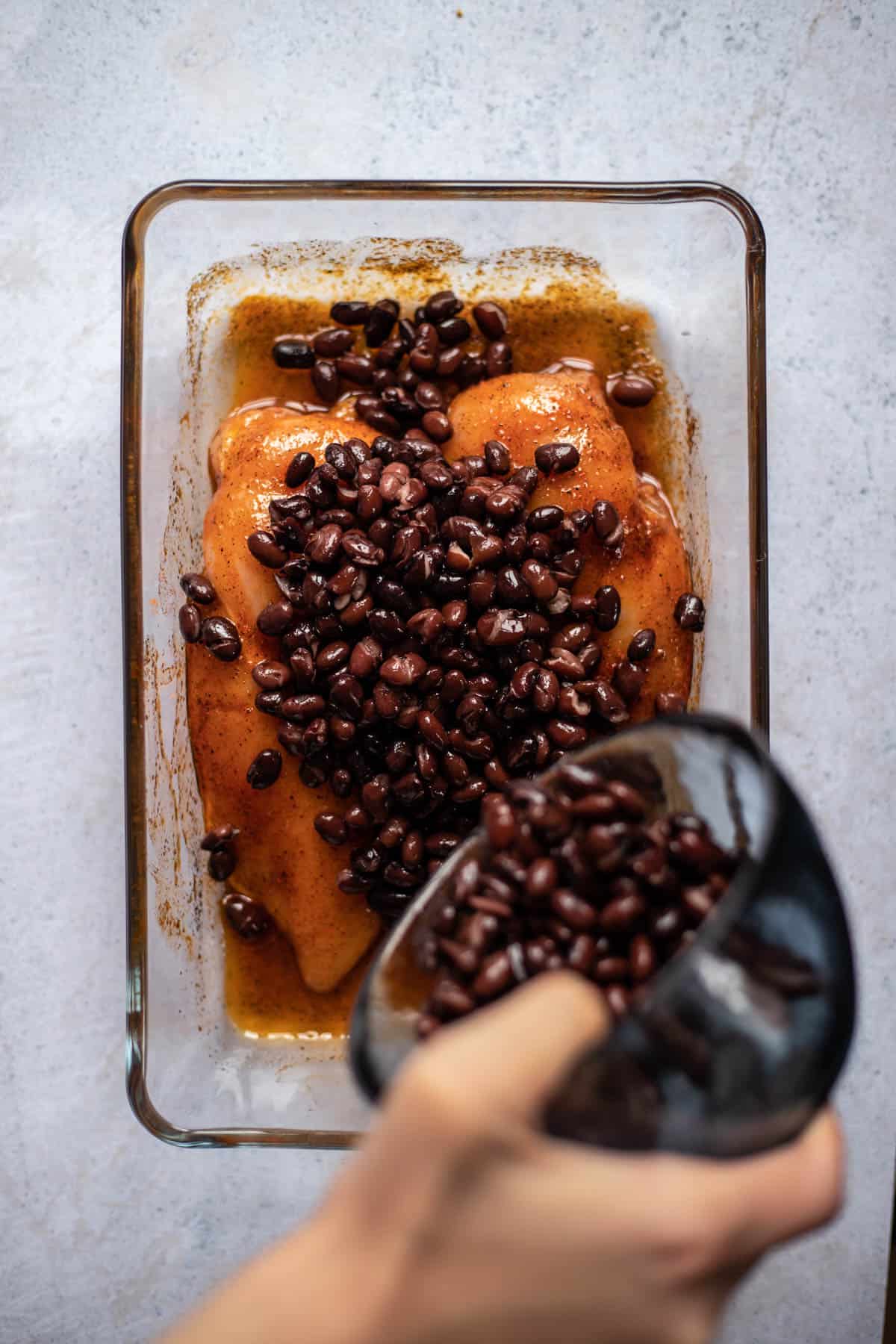 black beans being poured over raw seasoned chicken breasts in a baking dish