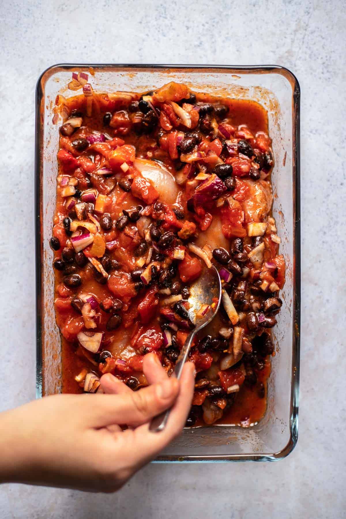 a hand using a spoon to stir salsa, beans, and red onions over two raw chicken breasts
