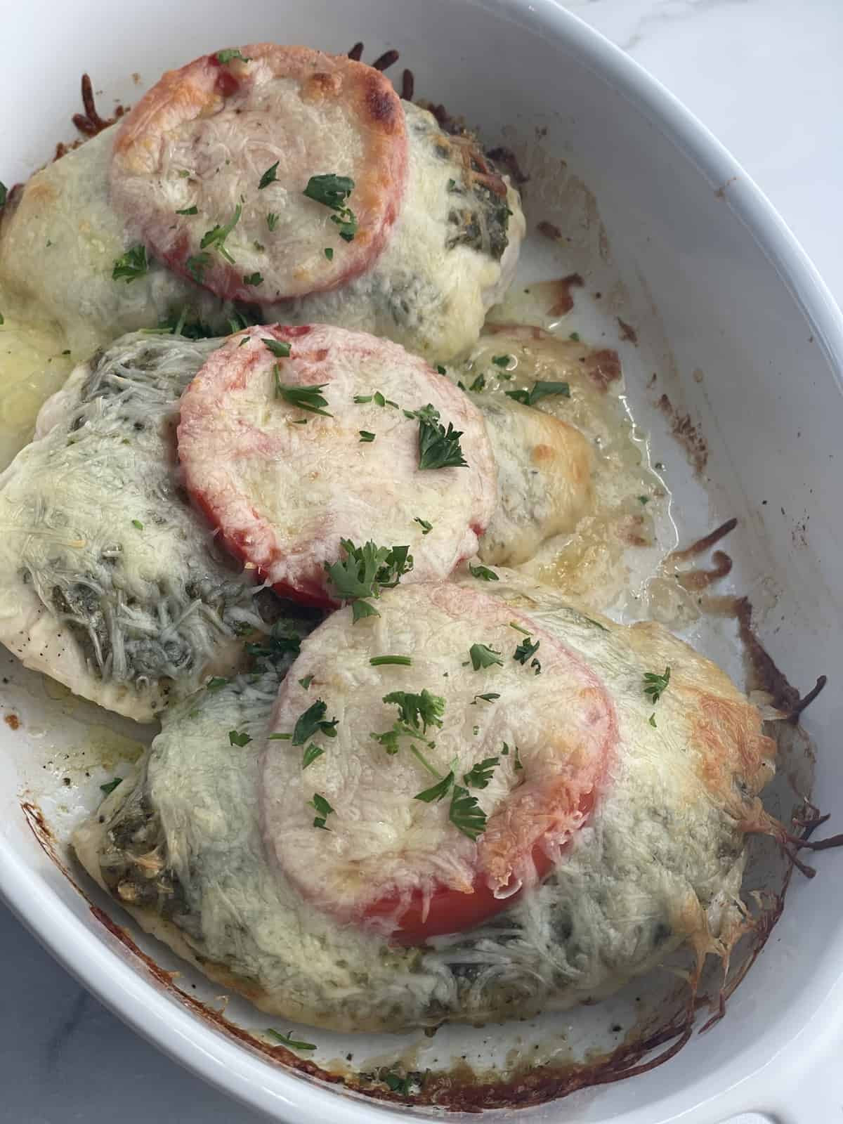 a white baking dish full of three chicken breasts topped with cheese, pesto, tomato slices, and herbs