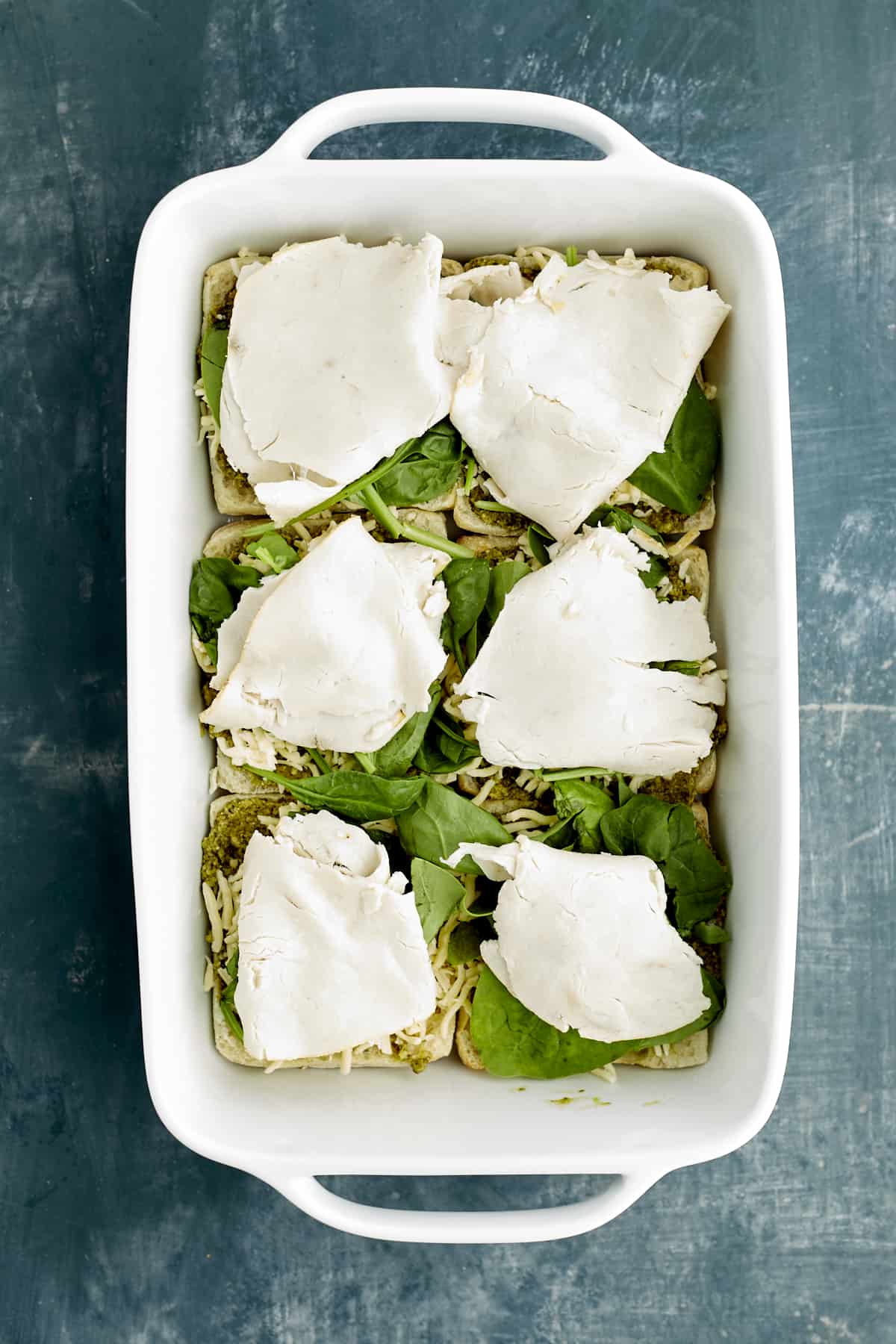 sliced ciabatta buns in a white baking dish layered with pesto, spinach, and turkey.
