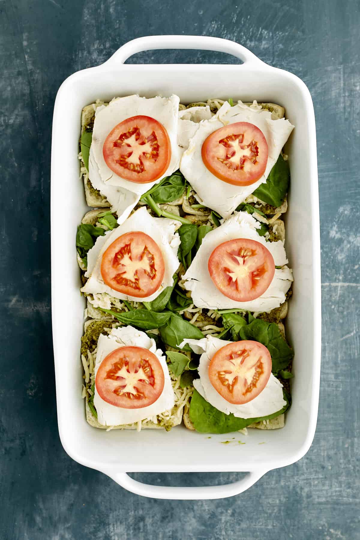Sliced ciabatta buns in a white baking dish layered with pesto, spinach, turkey, and tomatoes
