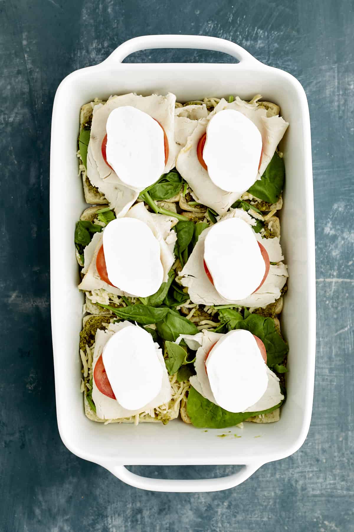 sliced ciabatta buns in a baking dish layered with pesto, spinach, tomatoes, turkey, and mozzarella.
