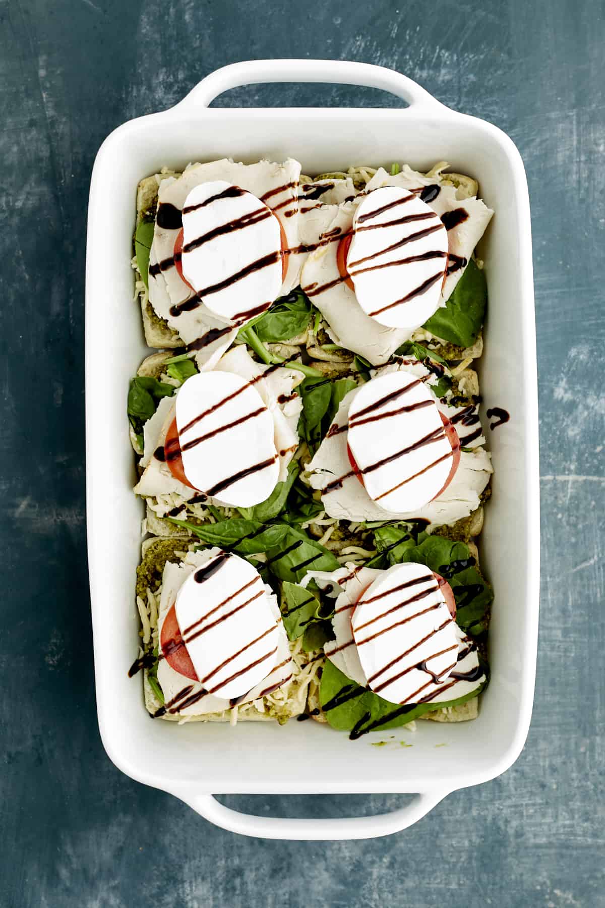 sliced ciabatta buns in a white baking dish topped with pesto, spinach, turkey, tomatoes, mozzarella, and balsamic glaze.