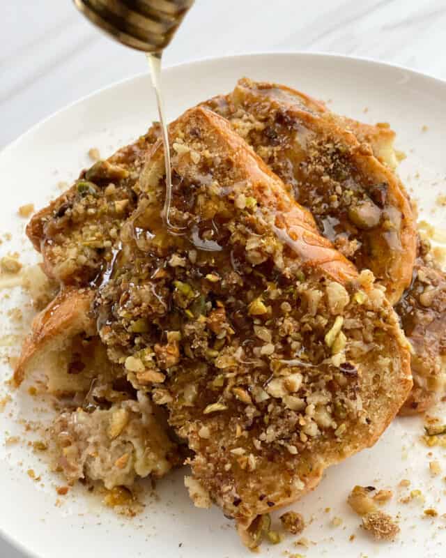 baked baklava brioche french toast topped with chopped walnuts, pistachios, and honey on a white plate