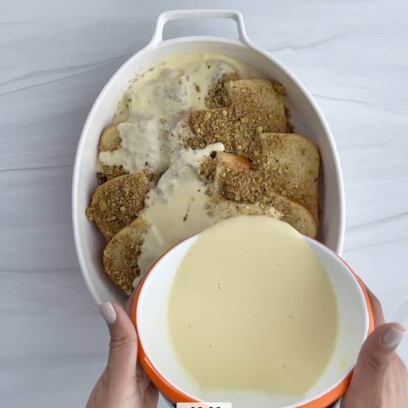 pieces of brioche bread topped with cinnamon sugar and chopped nuts with two hands pouring a creamy custard mix on top
