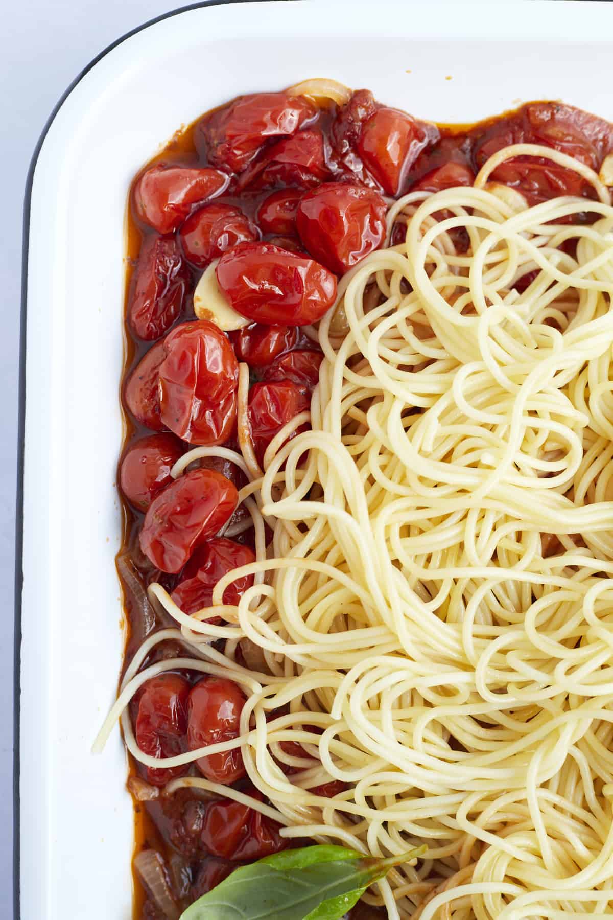 Cooked spaghetti noodles on top of roasted tomatoes, garlic cloves, and onions in a baking dish
