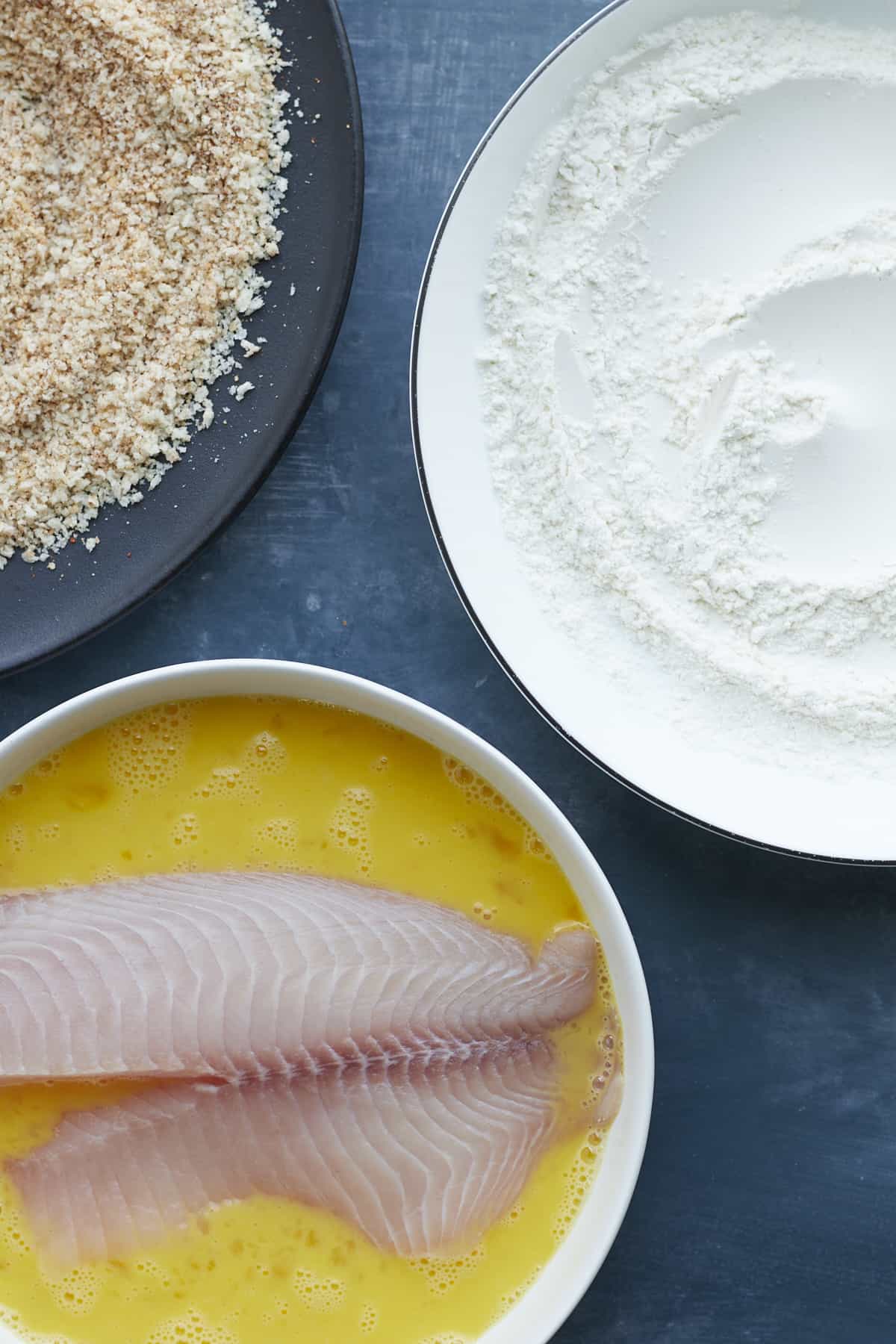 tilapia being dredge in egg wash with flour and breadcrumbs to the side