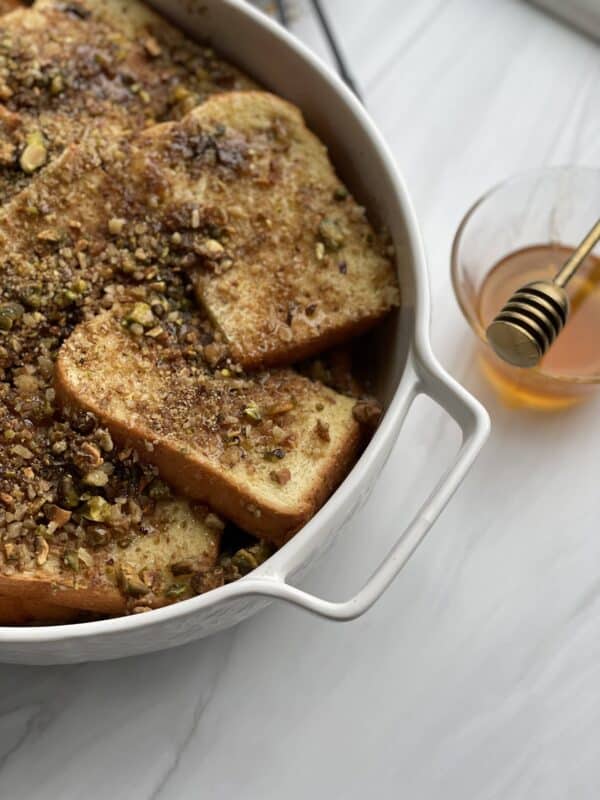 baked baklava brioche french toast in a white baking dish with a small bowl of honey on the side