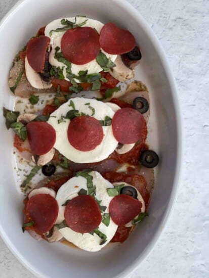 overhead image of three raw chicken breasts topped with pizza sauce, mozzarella, pepperoni, olives, and fresh herbs