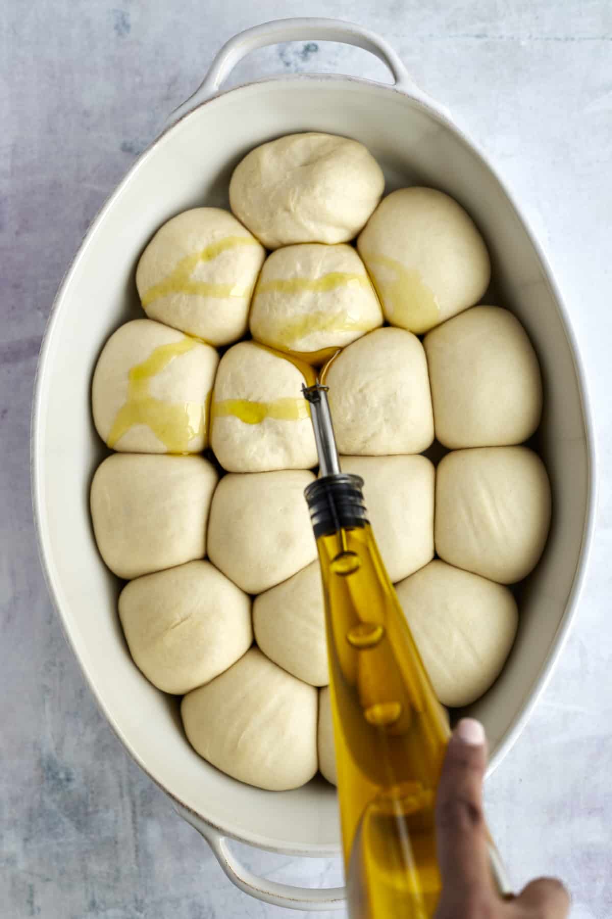 olive oil being drizzled over raw bread rolls in an oval baking dish 