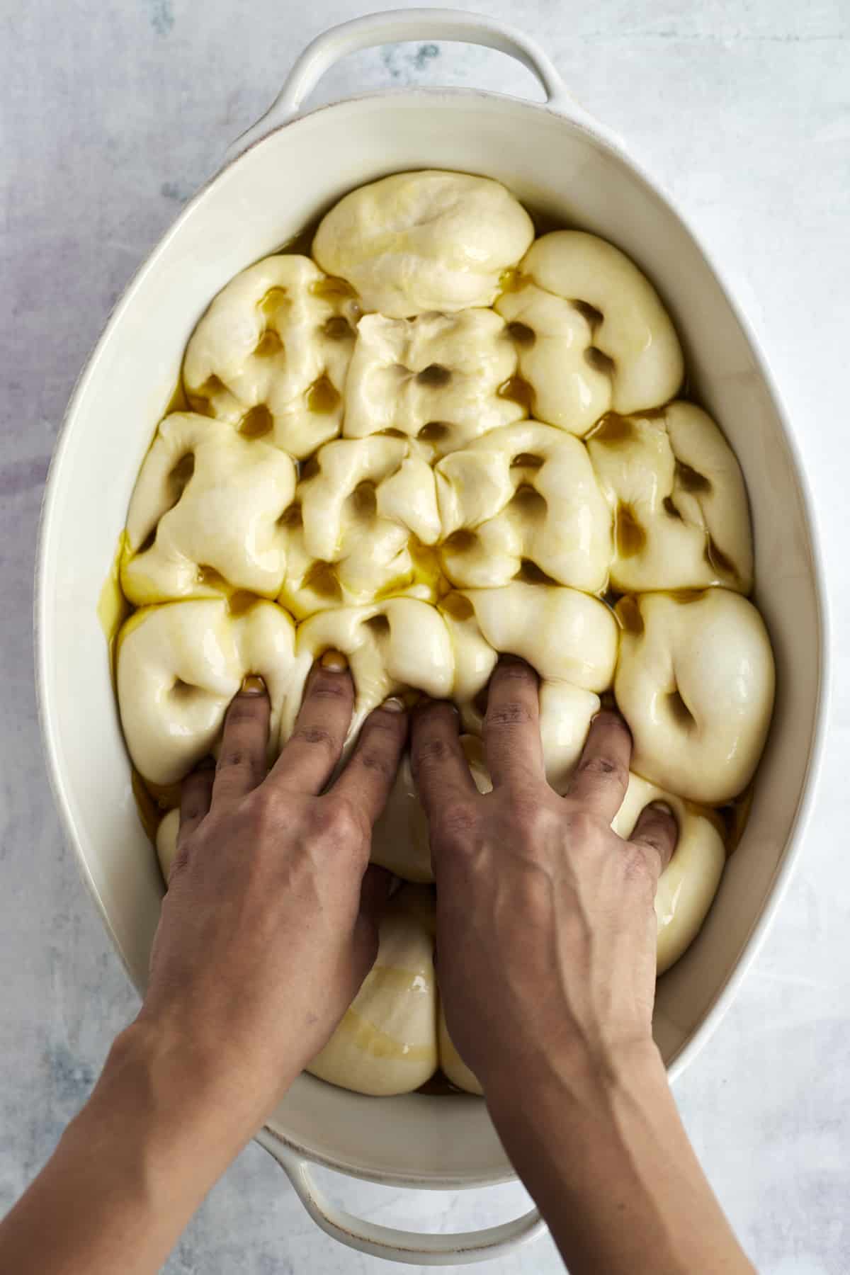 hands pressing indents into bread roll dough covered with olive oil 