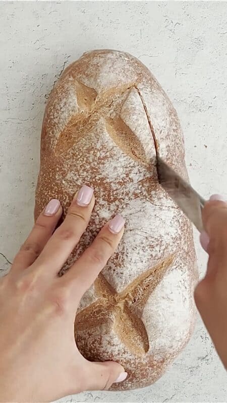 a woman's hands cutting the top off a loaf of sourdough