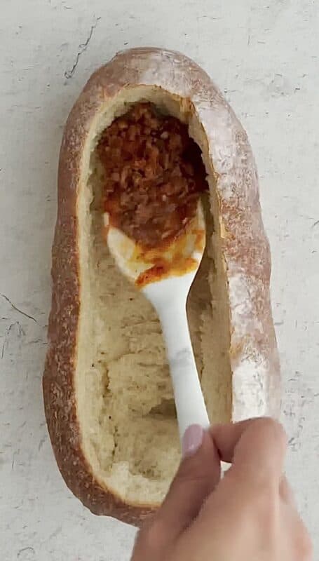 overhead image of a sourdough bread bowl with a woman's hand adding a spoonful of bolognese sauce in the bottom