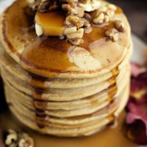 a stack of healthy pumpkin pancakes on a plate topped with a pad of butter and walnuts with maple syrup being drizzled on top