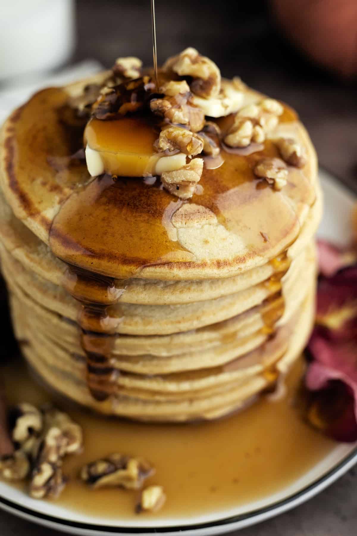 a stack of healthy pumpkin pancakes on a white plate topped with a pad of butter and walnuts with maple syrup being drizzled on top